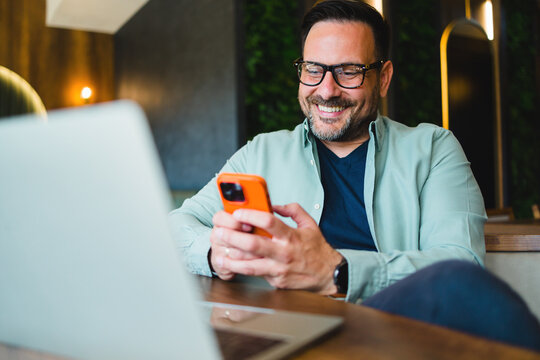 Smiling man using smartphone while sitting with laptop in cafe