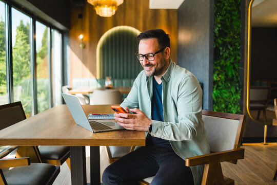 Smiling man using smartphone while working on laptop in cafe