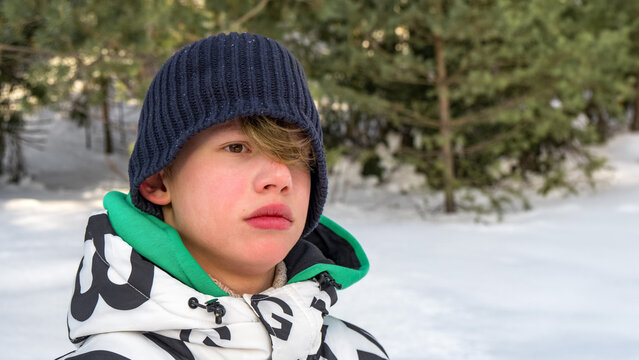 Portrait of teenager in winter forest. He wears thick jacket and knitted hat. His long hair is visible under beany. Snow covers ground and trees surround him