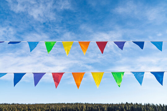 Bright flags in yellow, orange, green, blue, and purple hang against wispy clouds sky. Flags sway gently from line above forest, at local event in countryside