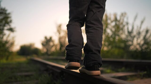 Child is walking on railroad track at sunset. Boy balances on rail in sneakers during summer evening. Legs step on train tracks outdoors. Kid walks away in nature with sun light in background.