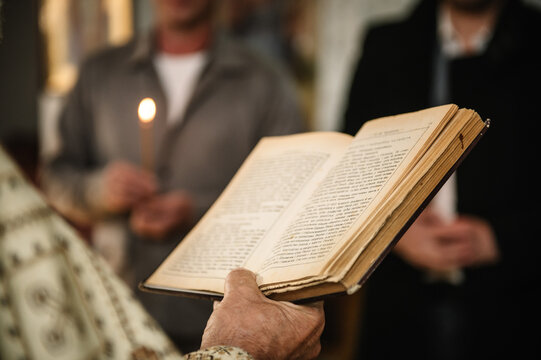 Close-up of an Orthodox priest's hand holding an open liturgical book during a religious ceremony. In the blurred background, people hold lit candles, creating a solemn and spiritual atmosphere.