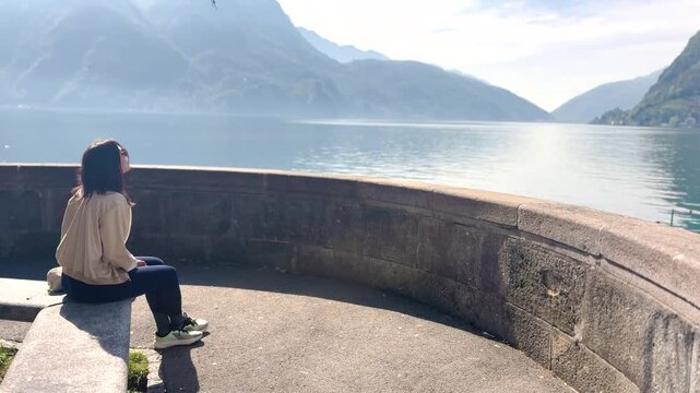 Young female tourist sitting at viewpoint overlooking Lake Lugano and Monte San Salvatore, enjoying scenic mountain landscape, peaceful travel moment in Switzerland