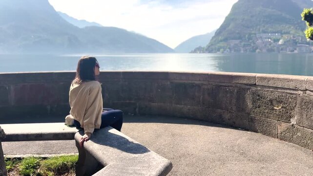 Young female tourist sitting at viewpoint overlooking Lake Lugano and Monte San Salvatore, enjoying scenic mountain landscape, peaceful travel moment in Switzerland