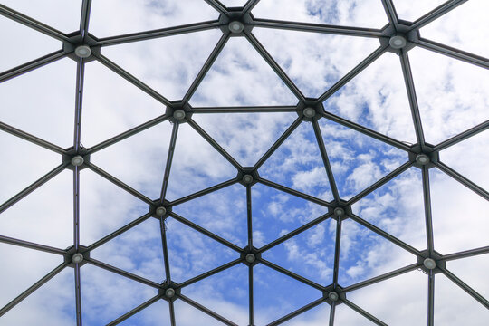 Steel geometric dome structure with interconnected metal nodes and repeating polygon patterns against a bright sky, highlighting architectural symmetry, engineered design and the visual rhythm of open