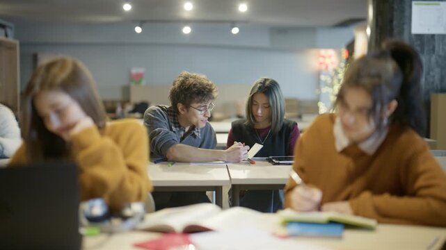 Young adults concentrating on their academic tasks and interacting thoughtfully at desks in a well-lit study hall.