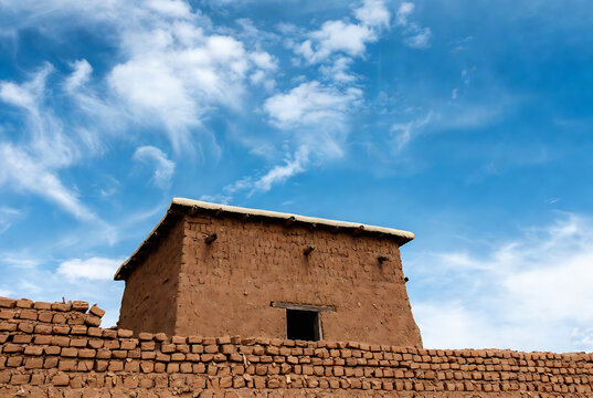 A clay adobe hut sits atop a brick wall under a vibrant blue sky with puffy clouds.