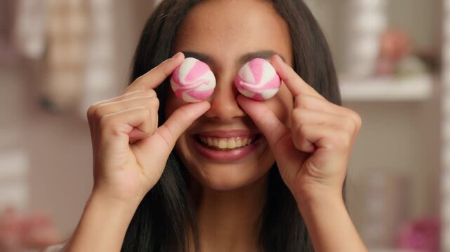 Happy smiling ethnic Latina girl holding two candy like glasses covering eyes grimacing cheerful Hispanic American woman female hold candies sweets marshmallows like eyeglasses smile in pink bathroom