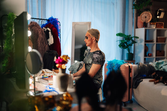 Drag performer sitting backstage, wearing wig cap and makeup, preparing for a show in a dressing room