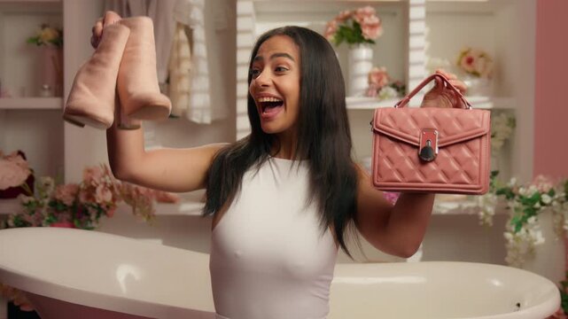 Pretty positive Hispanic girl Latina woman holding pink bag and high heels shoes boots preparing for date happy hold cloth accessories smile looking at camera American female in glamour candy bathroom