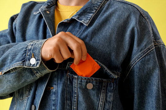 Young man putting condom in jeans jacket's pocket on yellow background, closeup