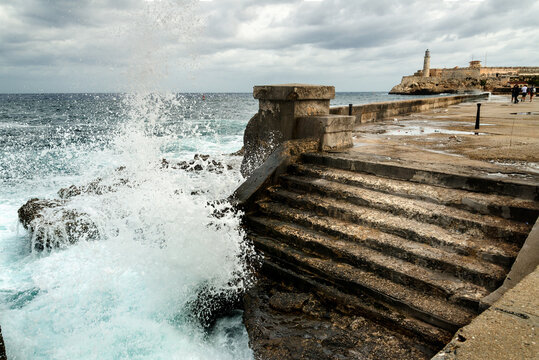 Cuba, Havana, The Malec&oacute;n.  Waves crash on the wall of the famous Avenida de Maceo, pedestrian walkway.  The Lighthouse of the El Morro castle in the background.  Faro Castillo del Morro