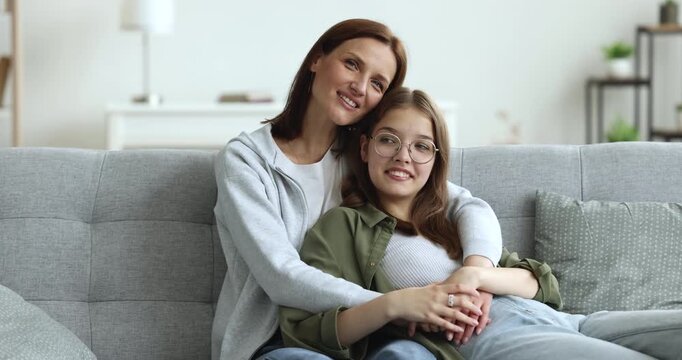 Loving mother embracing daughter, enjoying casual conversation, relaxing together on couch in living room at home. Family bond, intergenerational connection, understanding between teen and parent