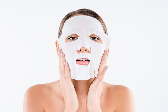 Beautiful young woman putting textile face mask on skin isolated on white background, cosmetic treatment, skincare routine, hydration care and spa wellness concept