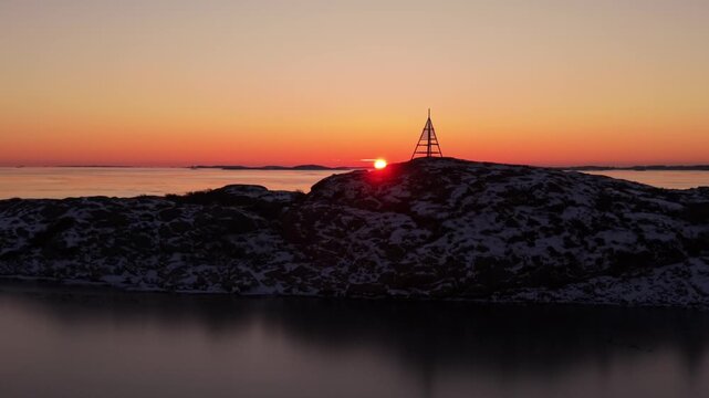 Aerial view of sunset over a rocky, snow-dusted outcrop with a wooden navigation mark silhouetted against the vibrant orange sky, Hovas, Vastra Gotalands lan, Sweden.