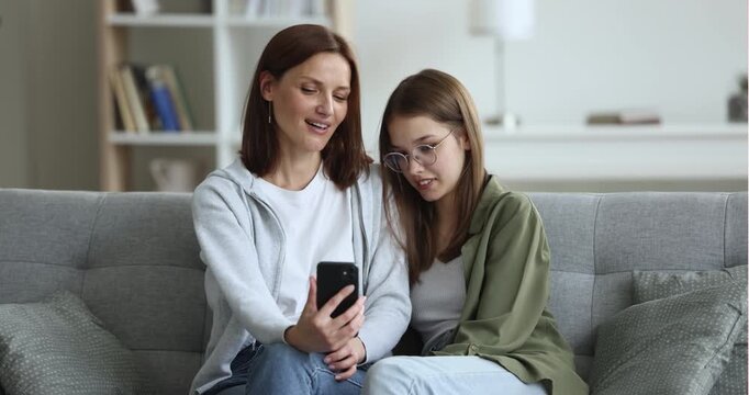 Mother and teen daughter sit together on sofa, having fun with mobile application, watching online content, posing for selfie using smartphone in home living room. Family bonding, social media use