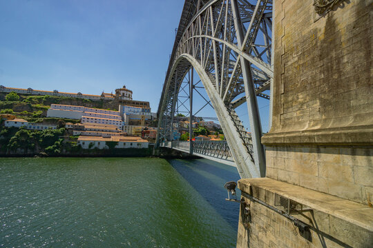 Porto, Portugal - 13 July 2021: View of the iconic Dom Luis I Bridge's metallic arch soaring over the shimmering Douro River, framed by the ancient city's weathered stone architecture.