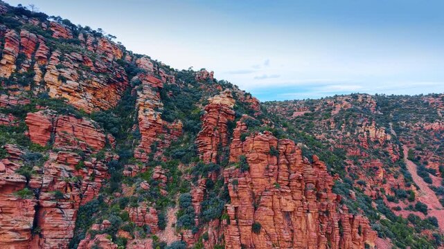 Textured red sandstone cliffs with green Mediterranean vegetation. Side view of layered rock formations and bushes under a cloudy blue sky.