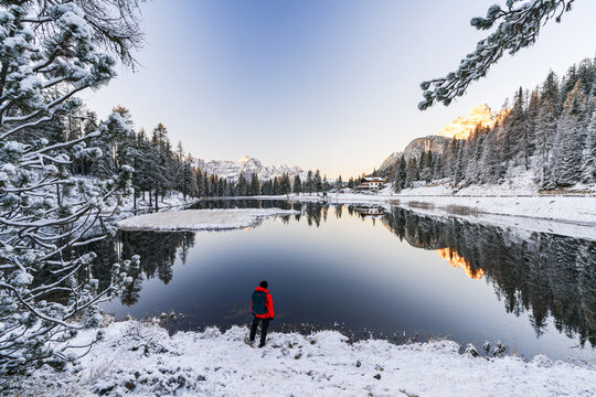 View of a solitary figure in a vibrant red jacket stands on the snowy shores of a serene lake mirroring the snow-dusted evergreens and distant, majestic mountains, Misurina, Veneto, Italy.