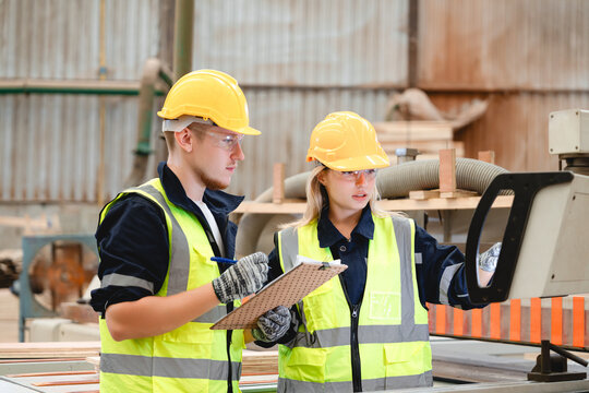 Two technicians wearing hard hats and safety vests meticulously inspect a large industrial machine and wooden planks within a dimly lit warehouse environment for quality assurance and maintenance.