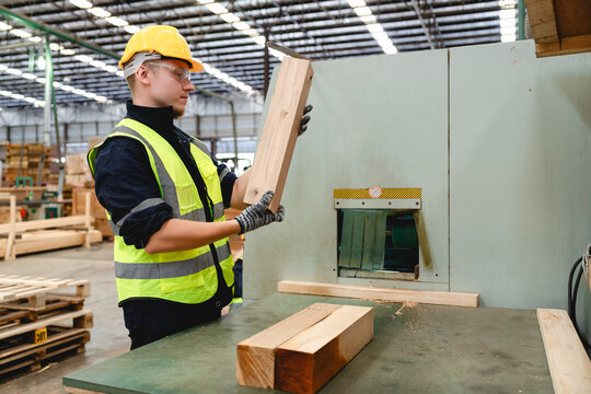 Caucasian worker operates a wood planer in a dimly lit factory, focused on precise wood processing while wearing a bright yellow safety vest and surrounded by wooden pallets.