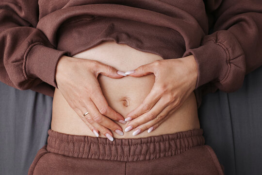 Woman forming a heart shape over her bare abdomen with both hands. Wellness and body awareness concept. Healthy lifestyle gesture focused on belly care, digestion and self compassion indoors.