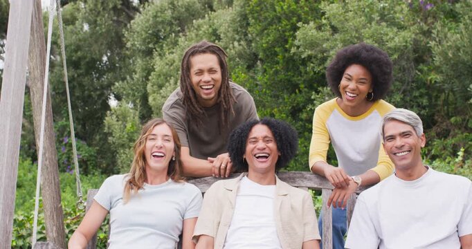Diverse friends laughing and leaning on wooden bench in leafy garden following camera prompt