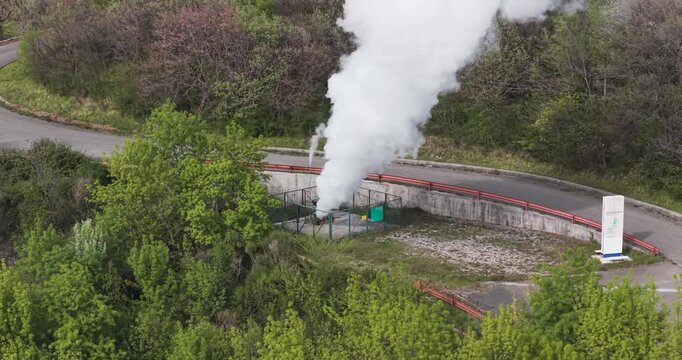 Larderello, Italy - 15 April 2026: Aerial view of Enel Green Power geothermal plant with a thick plume of white steam rising from a fenced wellhead surrounded by green trees and a road.