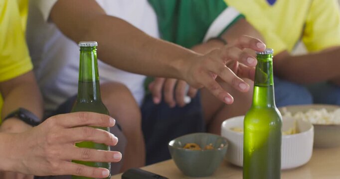 Diverse friends in jerseys reaching green beer bottles on couch, opening caps while watching match