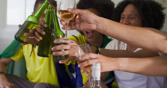 Diverse friends cheering after match goal, raising green beer bottles and wine glasses on couch