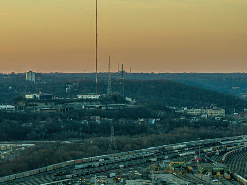 Cincinnati, United States - 27 February 2026: Aerial view of a train yard nestled amidst a dense forest under a warm, golden sky, with tall communication towers piercing the horizon.