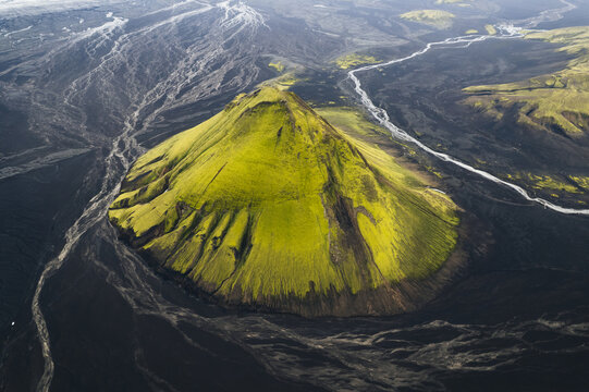 Aerial view of the vibrant green moss-covered Maelifell mountain starkly contrasts against the dark volcanic ash plains, crisscrossed by glacial rivers, Maelifell, Iceland.