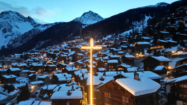 Aerial view of a brightly lit cross towering over snow-dusted chalets nestled in the valley, with majestic mountains embracing the twilight sky, Grimentz, Switzerland.