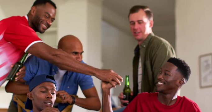 Diverse male friends gaming at home, bald friend tapping redshirted player, gamepads and beers