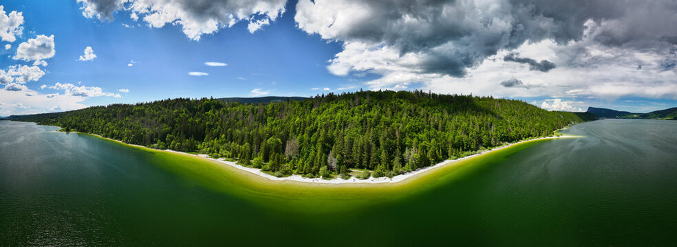 Aerial view of the vibrant green forest meeting the tranquil Lac de Joux under a sky of shifting clouds, Le Lieu, Vaud, Switzerland.