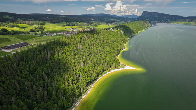 Aerial view of the tranquil lake mirroring the sky, bordered by verdant forests and rolling hills, a serene landscape of nature's artistry, Le Lieu, Vaud, Switzerland.