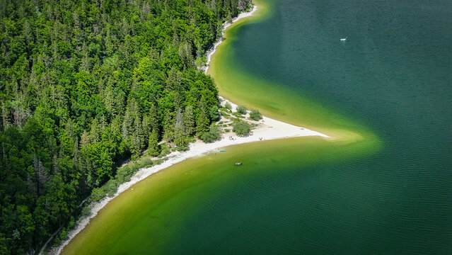 Aerial view of the lush forest meets the serene waters of Lac de Joux, where the interplay of light paints the sandy shores with hues of emerald, Le Lieu, Vaud, Switzerland.