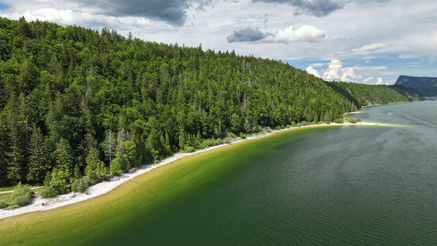 Aerial view of the serene Lac de Joux reflecting the verdant forests and dramatic cliffs under a sky of scattered clouds, Le Lieu, Vaud, Switzerland.