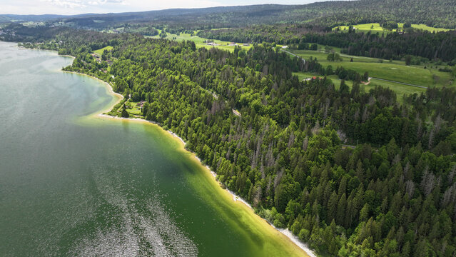 Aerial view of the serene Lac de Joux's shoreline, where emerald waters meet lush forests under a vast sky, Le Lieu, Vaud, Switzerland.