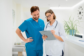 Fototapeta premium Male surgeon and young female doctor looking at clipboard with patient information in hospital corridor, medical teamwork, professional communication, healthcare staff collaboration and clinic service
