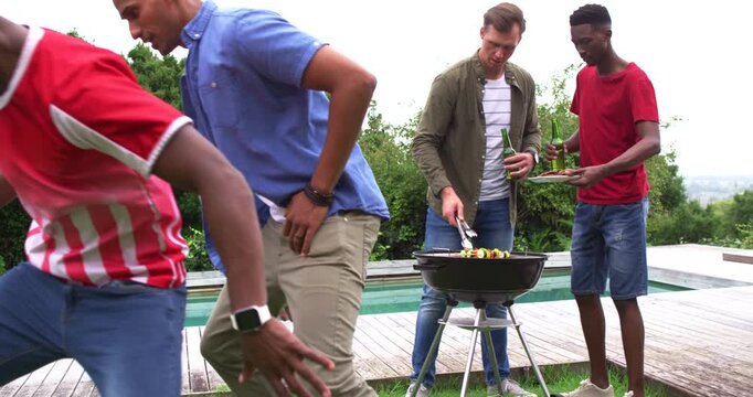 Diverse male friends, men running in, starting tussle for ball on lawn while cook turning kebabs