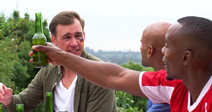 Diverse male friends clinking green bottles at yard after man with German facepaint offering toast