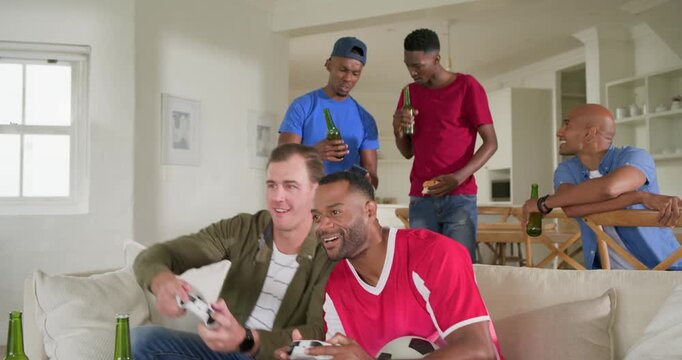 Diverse male friends arriving home, offering drinks, rooting and watching two men playing gamepads
