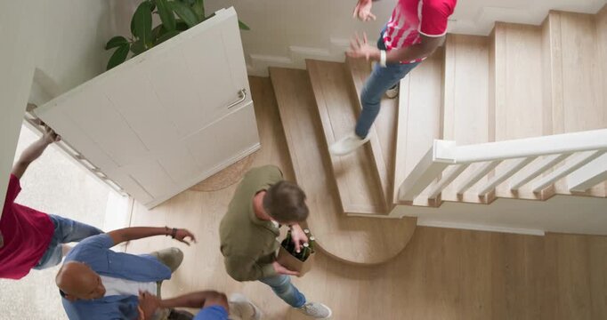 Diverse men entering to visit as door opening greeting, carrying plant and soccer ball in hall