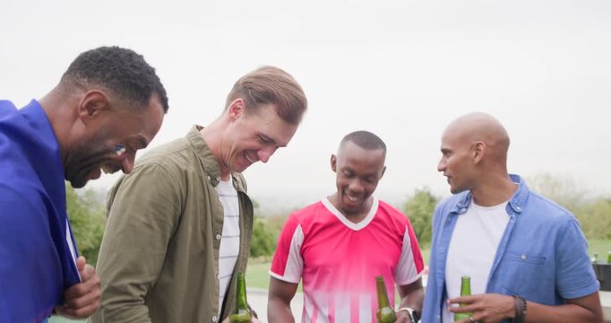 Diverse male friends grilling, sipping beer and kicking soccer ball on deck while watching match