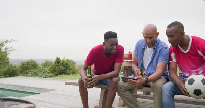 Diverse male friends watching tablet showing match goal on deck by grill and pool, clinking beers