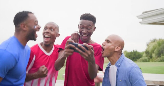 African American men leaning around phone after seeing match result, cheering and grilling on deck