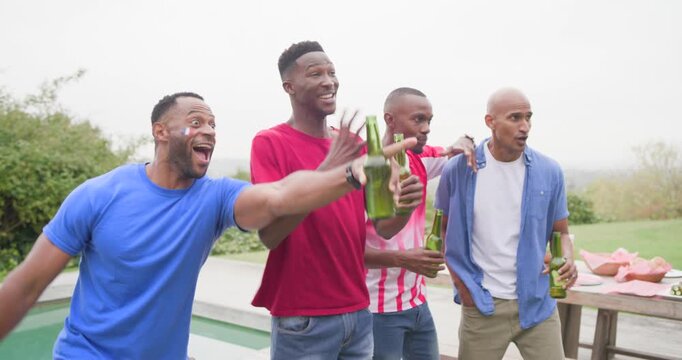 African American men reacting to off-camera call while celebrating, holding bottles on patio