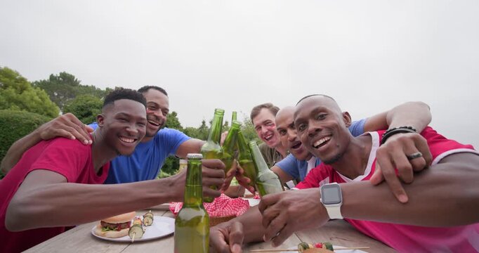 Diverse male friends leaning at picnic table, man reaching camera sparking toast with green bottles