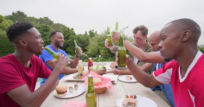 Diverse male friends raising bottles, clinking toast to start bbq, passing salad on backyard deck